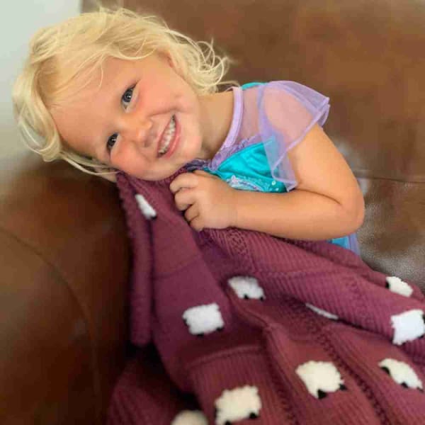 Young girl sitting with her raspberry maroon handmade baby blanket with 24 fluffy white sheep