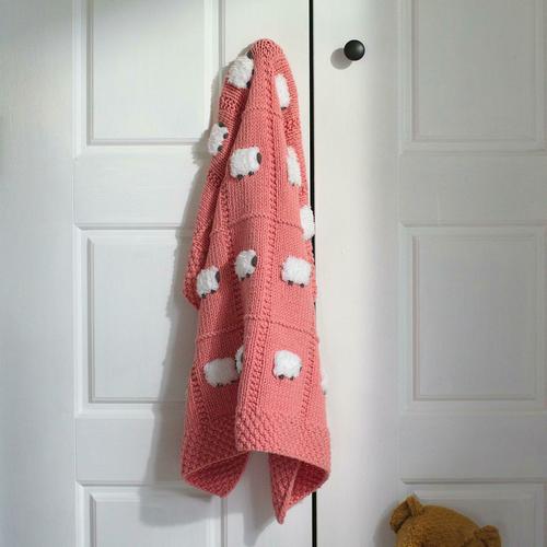 Salmon pink baby blanket hanging from a closet doorknob. The blanket has fluffy white sheep and is all knitted by hand.